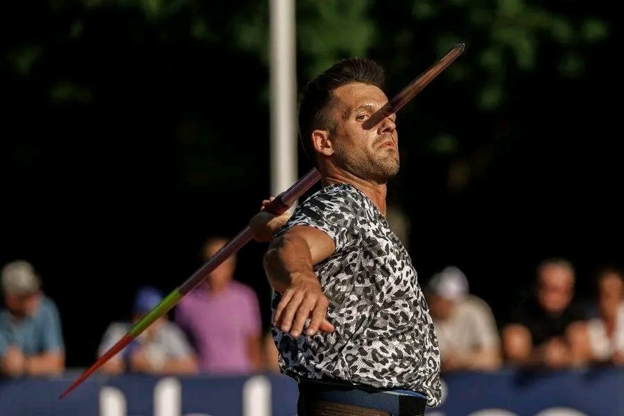 Športovec v akcii pri hode oštepom na atletickej súťaži, s publikom v pozadí.