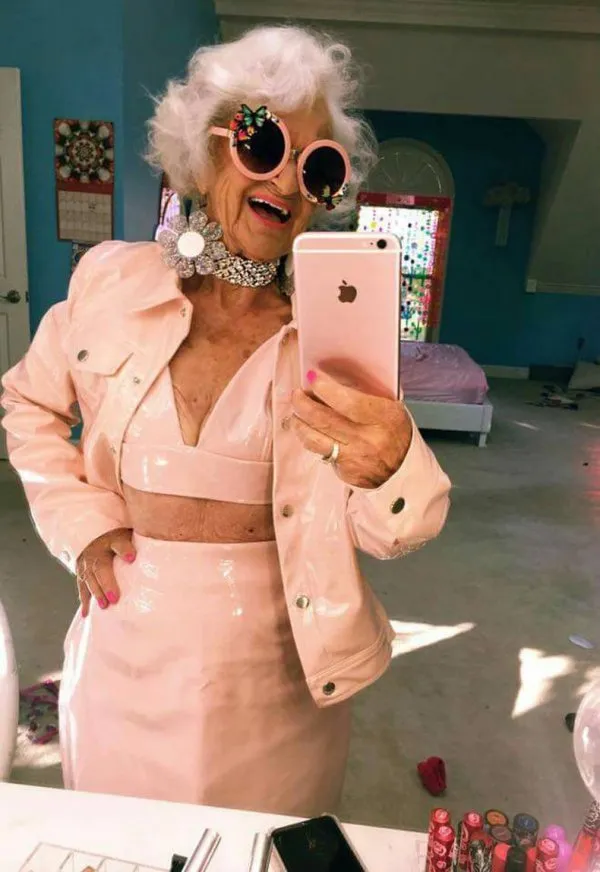 Štýlová staršia žena v ružovom oblečení a okuliaroch robí selfie v svetlej izbe.