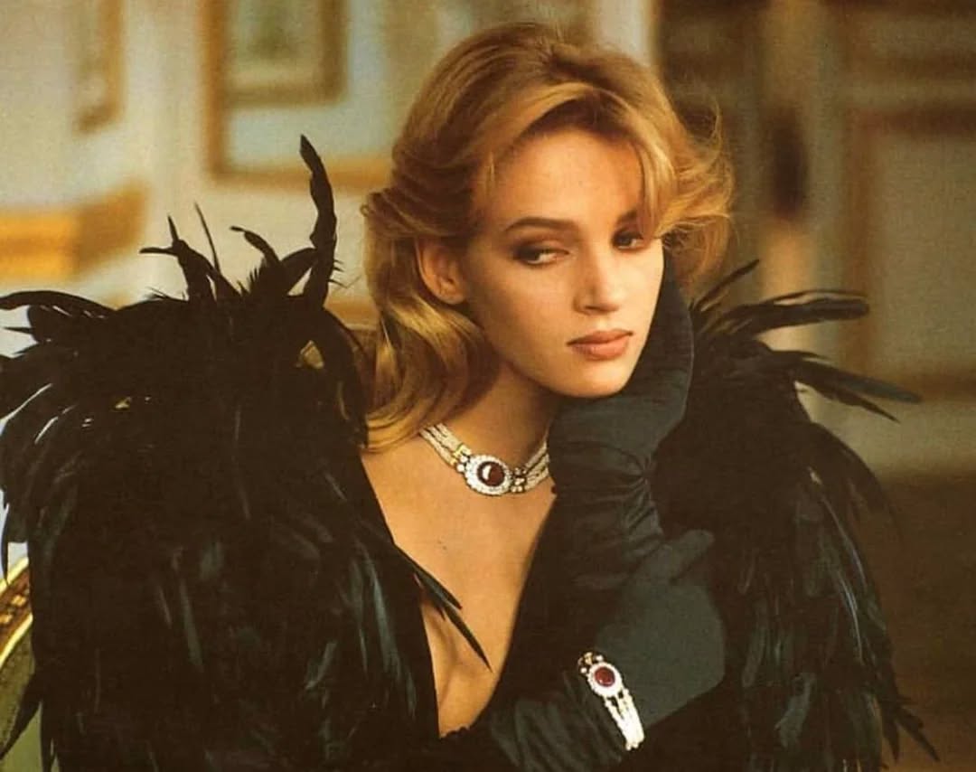 Žena v čiernej róbe s perím a šperkami, sedí zamyslená s rukou na tvári vo vnútri elegantnej miestnosti.