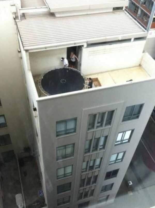 Trampolína na streche budovy s dvoma osobami stojacimi vedľa, pohľad zhora na mestské prostredie.