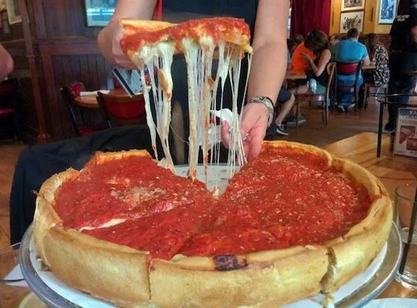 Osoba drží kúsok syrovej Chicago-style pizze s paradajkovou omáčkou, tavený syr sa hojne natiahne.
