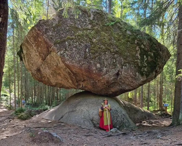 Žena v tradičnom odeve stojí pod obrovským balvanom v lese.