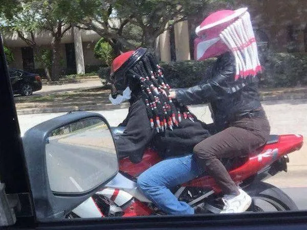 Dvaja ľudia na motorke s prilbami v tvare kreslených postavičiek, jazdiaci po ceste, príroda v pozadí.