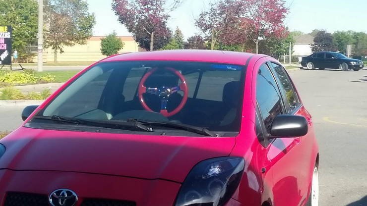 Červené auto s neštandardným volantom na parkovisku, stromy a ďalšie auto v pozadí.