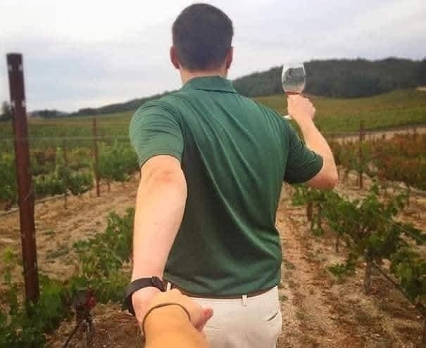 Osoba drží ruku partnera a kráča vinicou s pohárom vína, všade okolo sú viniče.