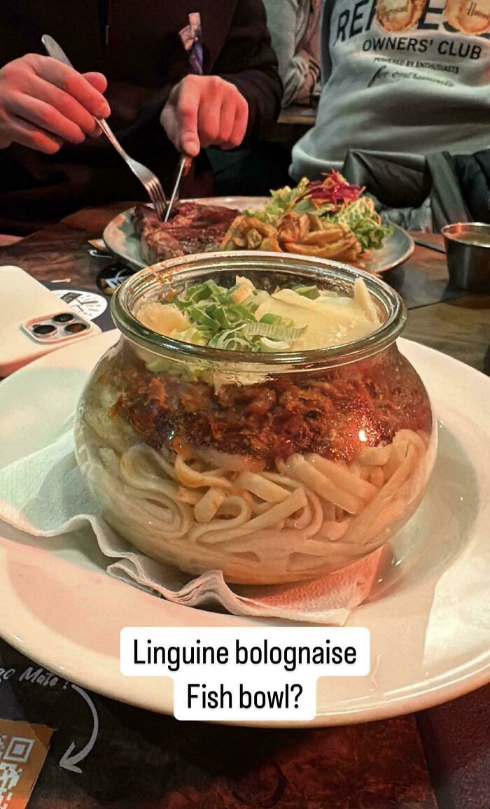 Sklenená miska plná linguine s bolonskou omáčkou a syrom v reštaurácii, vedľa taniera so steakom a šalátom.