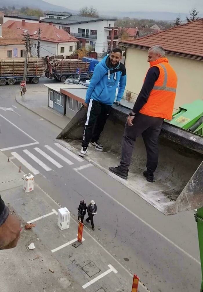 Dvaja muži stoja v lyžici bagra vysoko nad ulicou v meste, pričom nižšie sú vidieť kráčajúci ľudia.
