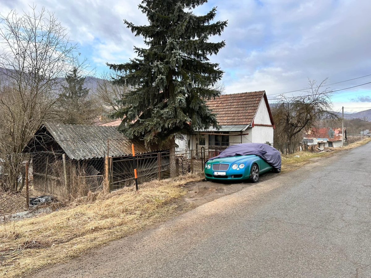 Luxusné auto pod plachtou pri ceste, vedľa tradičnej dedinskej chalupy na vidieku.
