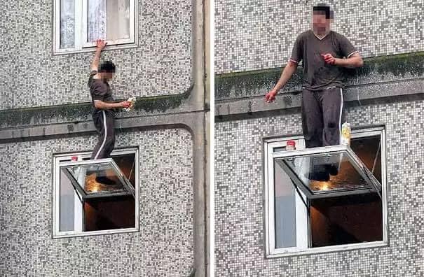 Muž stojí na vyklopenom okne výškového domu, bez viditeľného zabezpečenia, držiac handričku a čistiaci prostriedok.