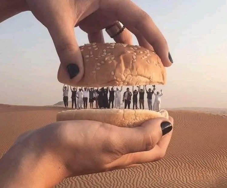 Obrovský hamburger držaný dvoma rukami nad púštnym pieskom; ľudia medzi vrstvami vtipne simulujú náplň.