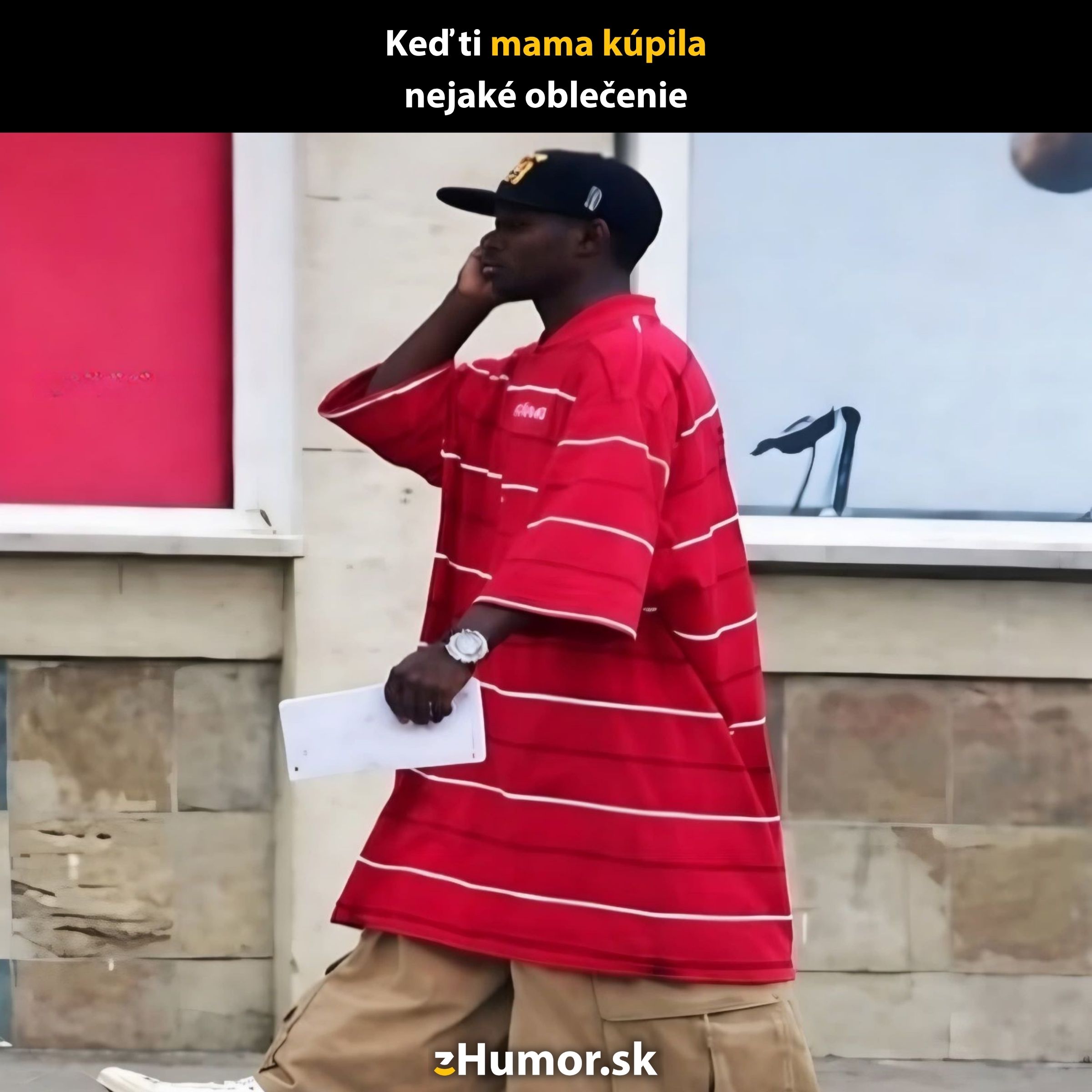 Muž v oversized červenom tričku kráča popri múre a telefonuje, oblečenie je komicky veľké.