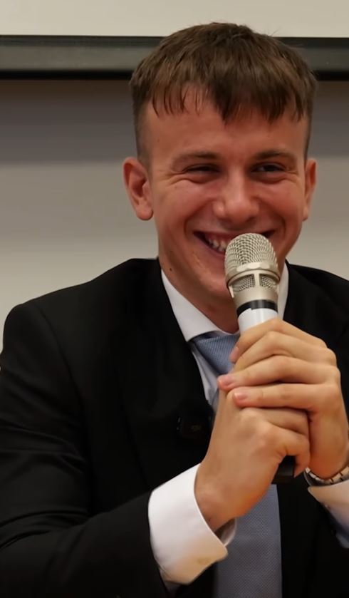 Muž v obleku usmievajúci sa s mikrofónom počas prejavu na konferencii.