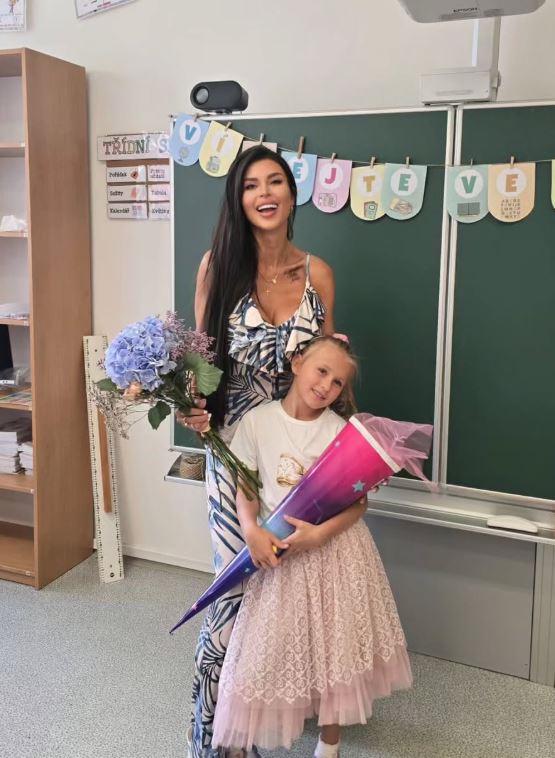 Učiteľka s dievčaťom v triede, kvety a školský kužeľ, uvítací banner na tabuli.